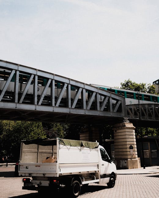 A white flatbed truck with a green cover over its cargo area is parked on a paved surface beneath a large metal railway bridge. The truck appears to be used for waste removal or delivery, with some cardboard visible inside the covered section. The bridge above is constructed from steel beams with a grey finish, supported by sturdy stone pillars, and supports an elevated train track, possibly part of an urban transit system. In the background, greenery from trees is visible, and the sky above is mostly clear with soft light indicating daytime. The scene suggests an environment centered around logistics or on-site clearance, with the presence of the truck implying alternative waste handling methods, potentially linked to private rubbish removal services by companies like Rubbish Removal Stockwell. The image reflects a scene of urban infrastructure combined with waste management activity, suitable for illustrating rubbish removal and private waste disposal services near public transit routes.