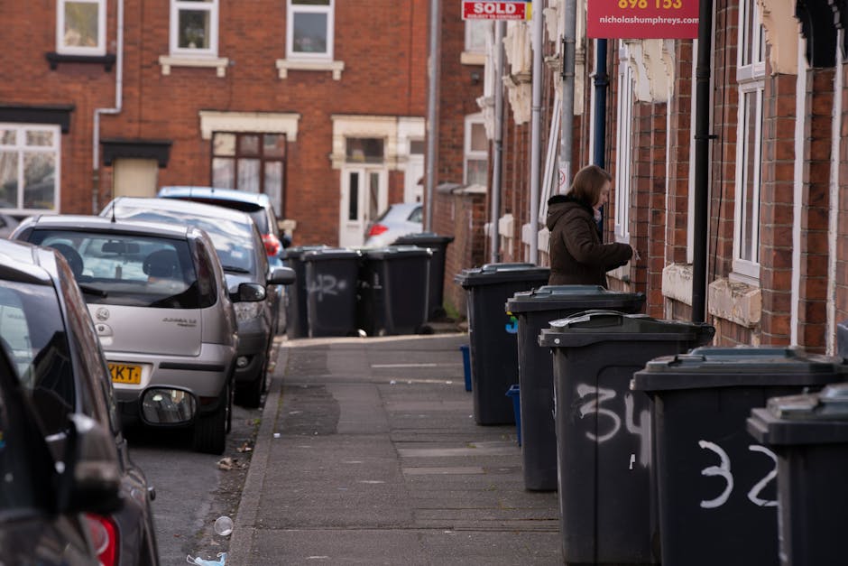 A woman in a dark brown jacket stands on a narrow sidewalk alongside a row of large black wheelie bins with white painted numbers, situated against the red brick exterior wall of terraced residential houses with white window frames. Some of the bins have partially open lids. The street is lined with parked cars of various makes and models, including a silver hatchback and dark-colored vehicles, occupying the curbside space. The pavement appears weathered with patches and scattered litter, while the background features more houses with similar architecture, including a 'SOLD' sign with red and white lettering attached to the wall, indicating a property sale. The scene reflects an urban setting with residents possibly preparing waste for collection or private disposal, aligning with practices of independent rubbish removal services such as those provided by Rubbish Removal Stockwell.