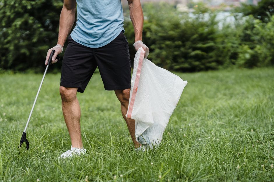 A person standing outdoors on a grassy lawn, wearing a light blue T-shirt, black shorts, and white sneakers, is engaged in removing garden litter. The individual is holding a large white biodegradable garden waste bag with a pinkish-color lining in one hand, while the other hand grasps a gardening tool, likely a grabber or waste picker, with the handle extended downward. The grass around them appears lush and green, and behind them, there is a background of dense, leafy trees and shrubbery, indicating a garden or park setting. The scene is well-lit with natural daylight, showing the person in the process of gathering organic waste such as leaves or grass clippings, a common activity associated with garden maintenance and private waste handling services. This visual underscores the type of outdoor rubbish collection that might be organized independently or through professional waste removal services like Rubbish Removal Stockwell, emphasizing on-site clearance or garden waste disposal activities.